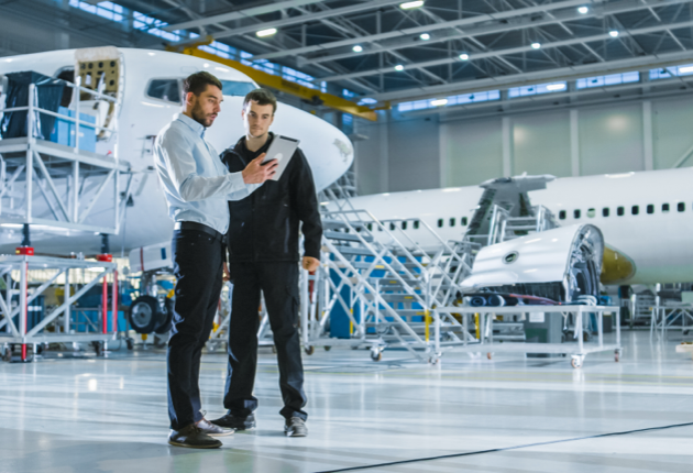 Two men standing in an airplane hangar, looking at a tablet.